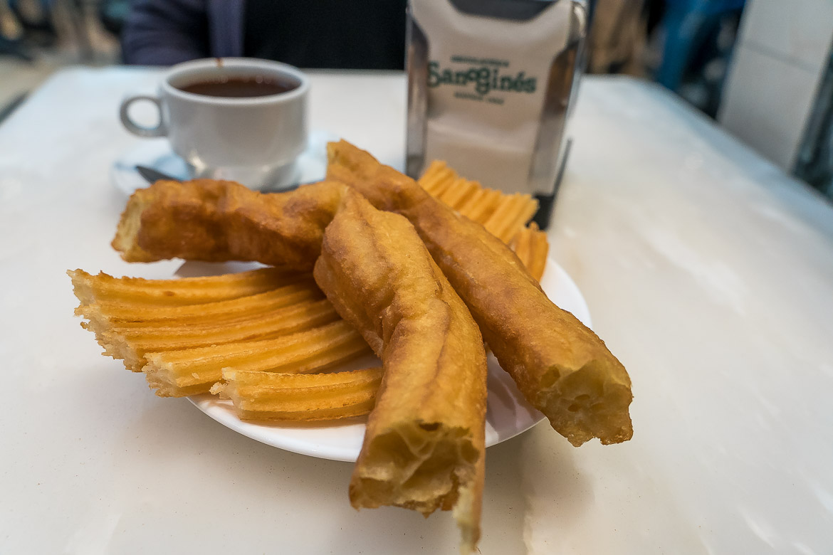 This image shows a plate with a portion of Churros and Purros in San Gines Chocolateria. In the background, there is a cup of chocolate.
