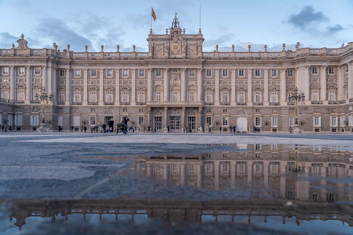 This image shows the main facade of the Royal Palace. The facade is reflected in a puddle of rain water in the foreground of the image.