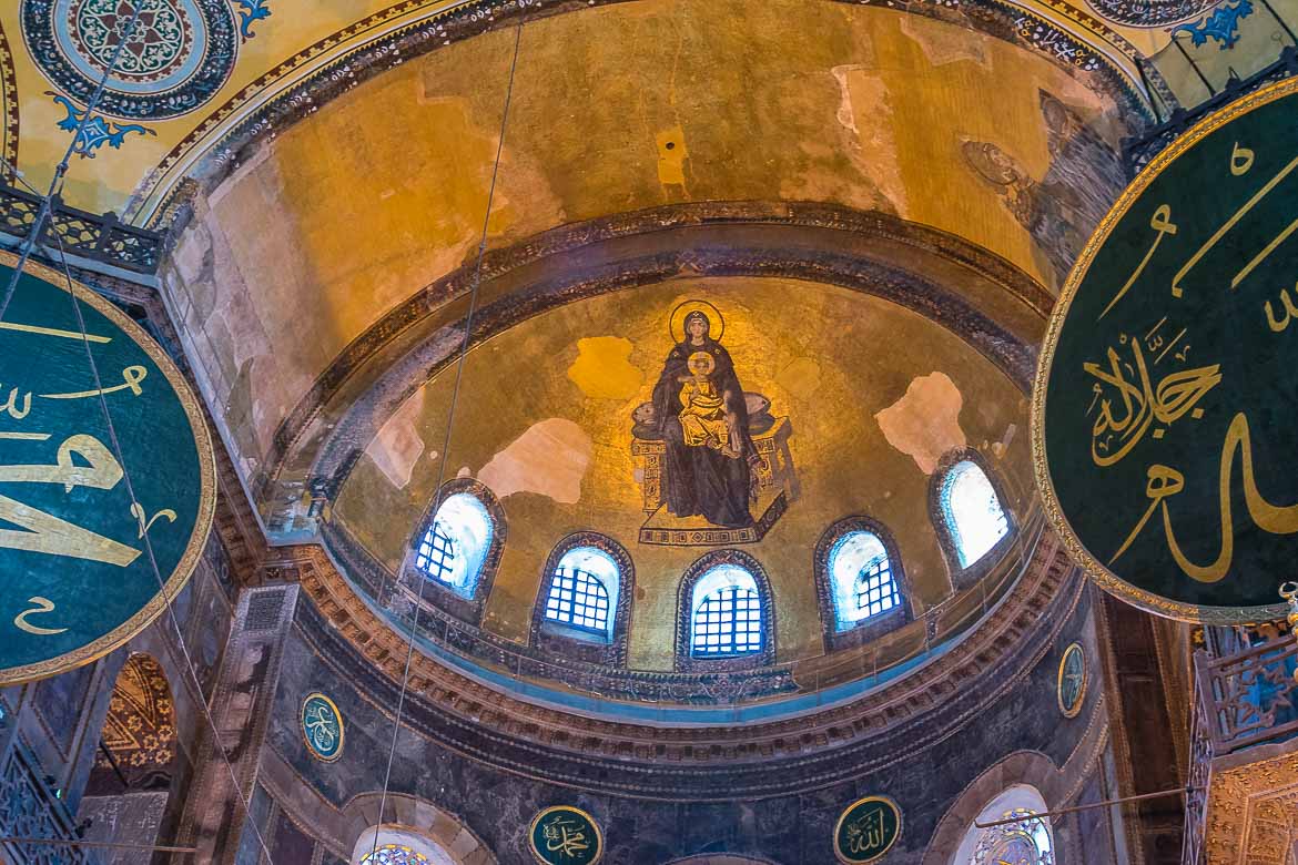A golden mosaic inside Hagia Sophia depicting Mary and Jesus. Left and right of the mosaic big round green signs with islamic texts  are hanging.