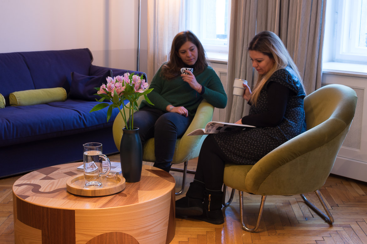 This image shows Katerina and Maria sitting comfortably at the living space of their hotel room reading a magazine and drinking tea. 