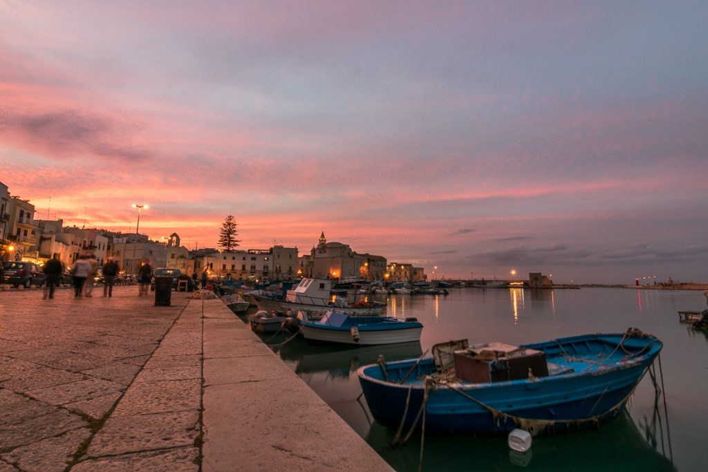 Best Beach Towns in Puglia Italy For Relaxing Holidays This image was shot in Trani port during sunset. The sky is a magnificent collection of pinks, yellows and blues, all reflecting on the mirror-like sea. There are small blue fishing boats in the sea and people walking along the promenade. The old-fashioned palazzi are dimly lit. We chose this photo to be the featured image for our article: Best beach towns in Puglia Italy for a laid-back vacation.