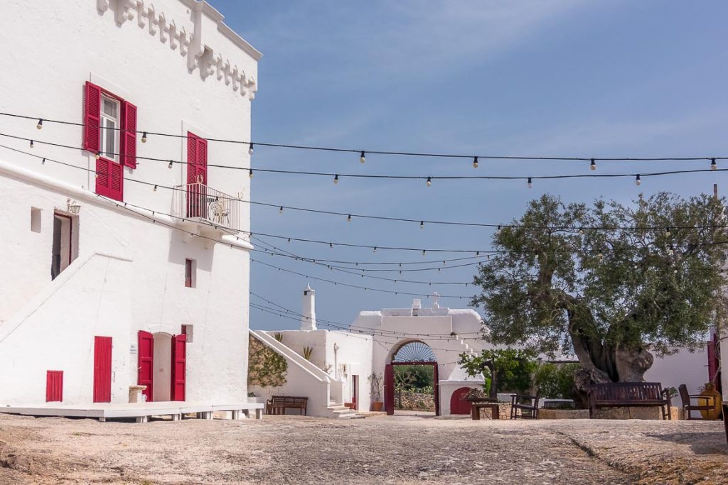 Best places to stay in Puglia Italy: An essential guide This photo shows the gorgeous central courtyard at Masseria Torre Coccaro. There is a whitewashed building on the left with bright red windows and doors. Masserie are typical to Puglia region. This is why we chose this photo to be the featured image of our article Best Places to stay in Puglia: An essential guide.