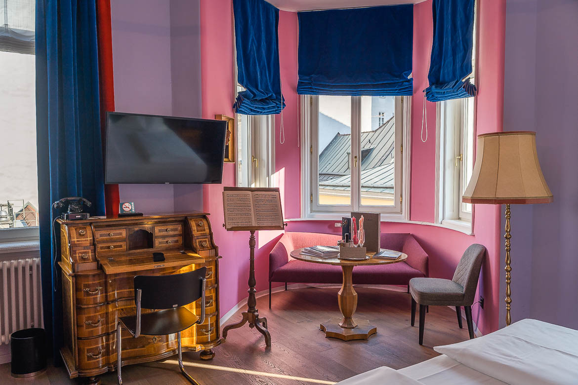 The small sitting area of our room at Beethoven Hotel. The room walls are pink and purple and the curtains on the windows are blue. There is also an antique desk with a TV over it.