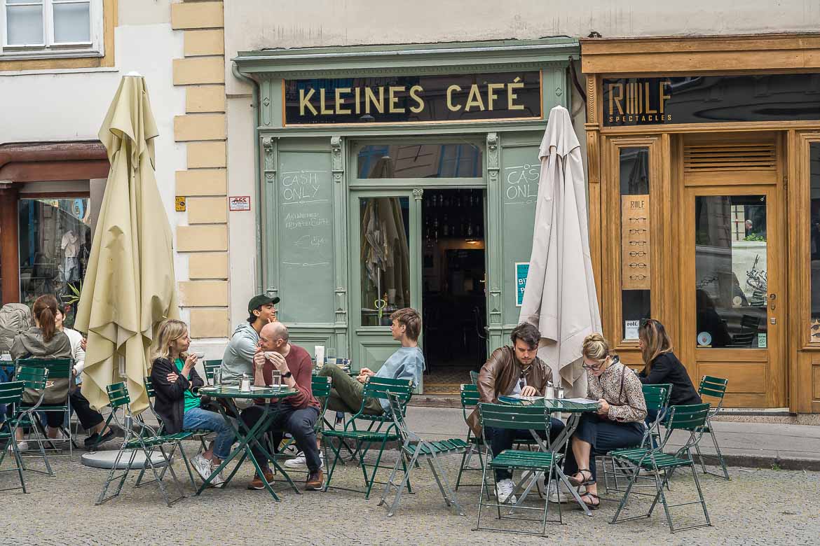 The outdoor seating area of Kleines Café. There are a few green tables with green chairs. The facade is also green with a big black sign with the name of the café.