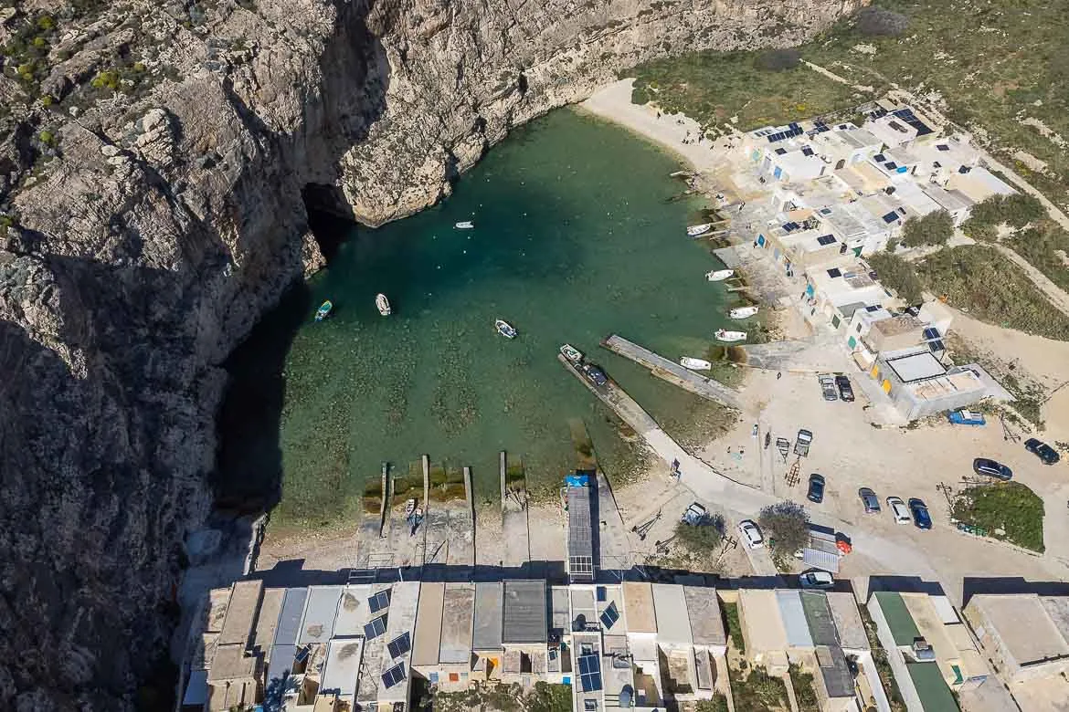 Panoramic view of Inland Sea in Gozo from a drone. Along the sea pool there are piers and fishermen sheds. Boats are departing from the pier and making their way to the tunnel in the rock leading to the sea. If you're wondering what to do in Gozo, exploring Inland Sea and around is a must. 
