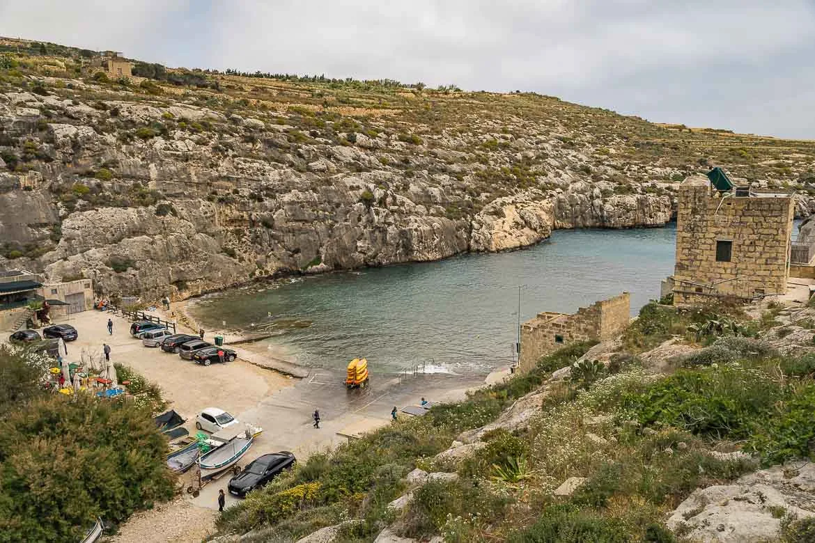 View of Mgarr Ix-Xini beach from the road atop the surrounding hill. There are a few cars parked near the beach and some canoes piled on the pier.