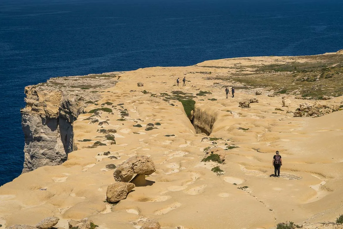 Panoramic view of the mushroom rock from the top of the surrounding rocks. A few rocks make the shape of a mushroom on the rugged plateau. Maria and a few people are walking around.