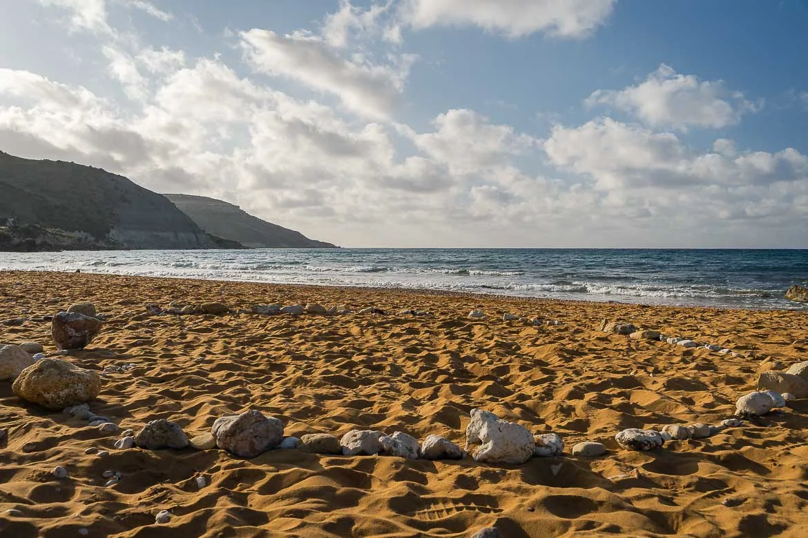 The red sandy beach of Ramla Bay from a low angle. A circle made of stones is formed on the red sand. If you're wondering what to do in Gozo, relaxing on the beach is a great idea. 