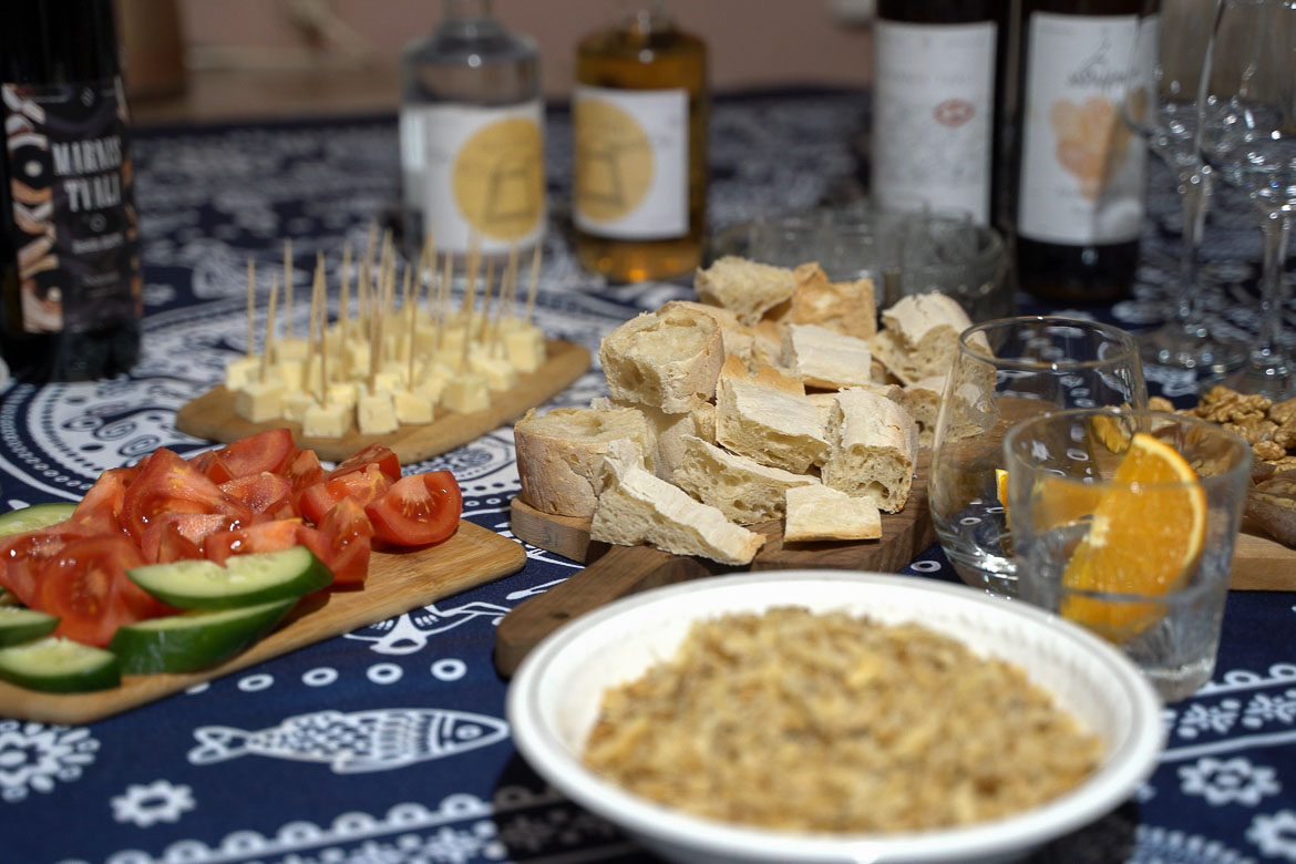 A table covered with a blue and white tablecloth. There's bread, tomatoes, cucumbers, cheese, wine and chacha on the table.