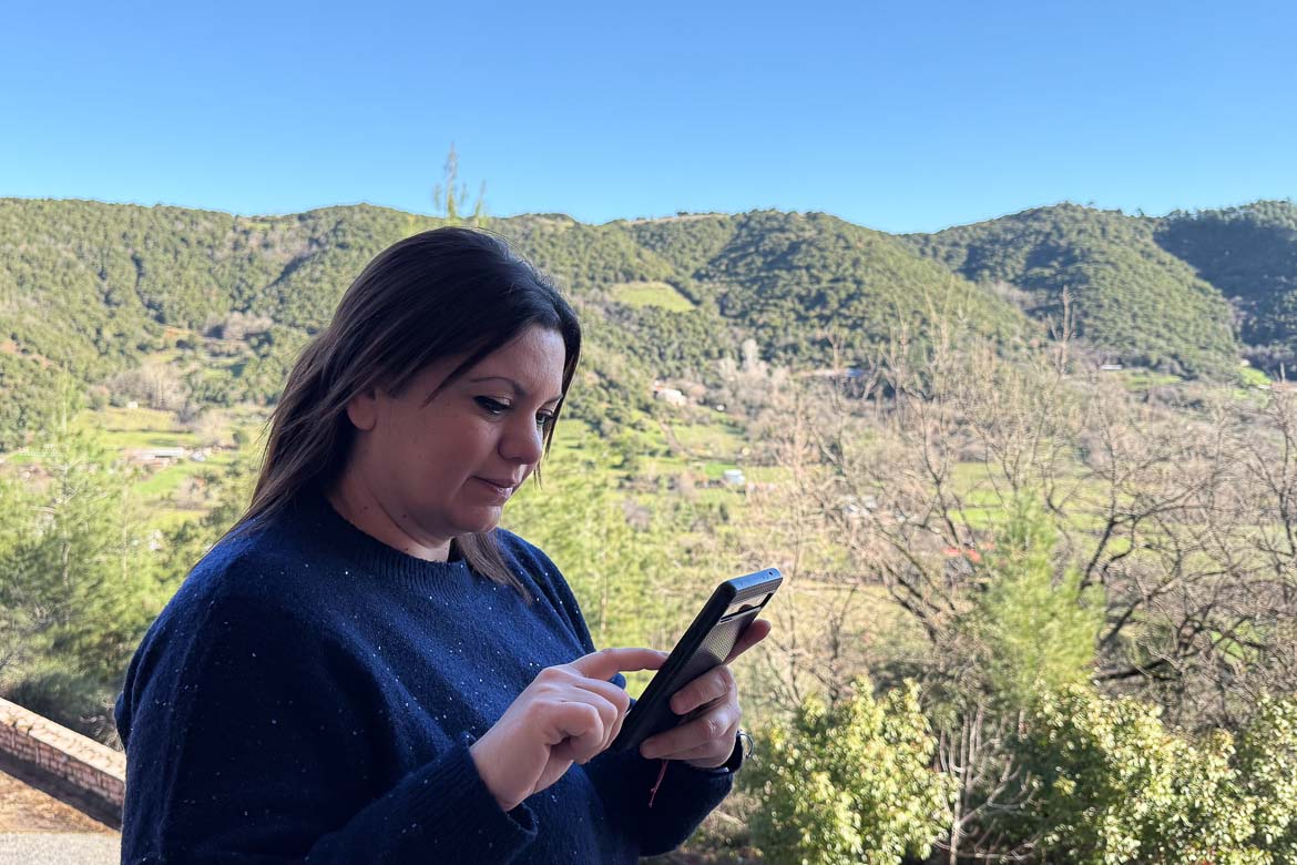 Maria in a blue sweater is scrolling on her cellphone. There are green mountains in the background.