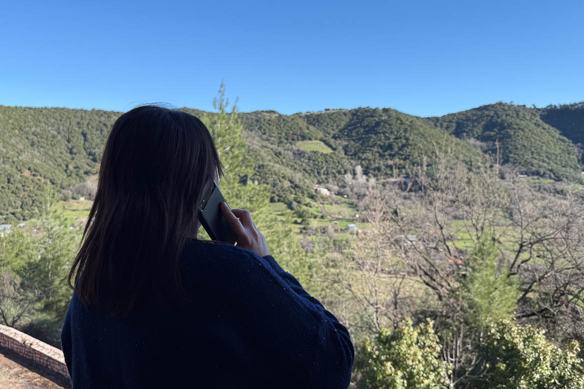 Maria with her back turned to the camera is talking on the phone. There are green mountains in the background.