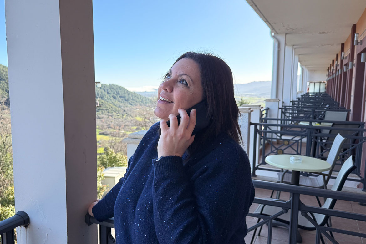Maria in a blue sweater is standing at a hotel balcony, looking upwards, talking on the phone and smiling.
