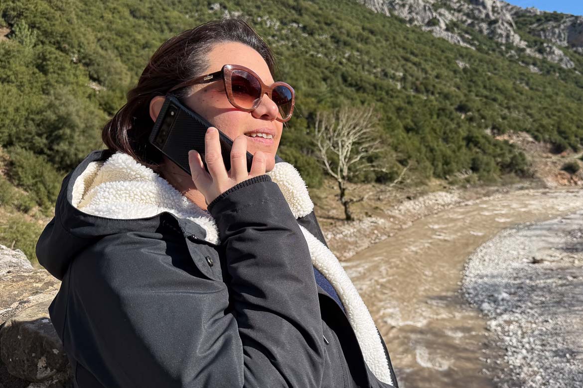 Maria in her winter coat and sunglasses is talking on the phone while standing on a bridge over a river.