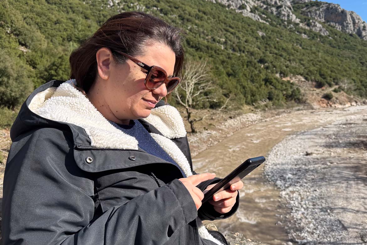 Maria in her winter coat and sunglasses is scrolling on her mobile phone while standing on a bridge over a river.
