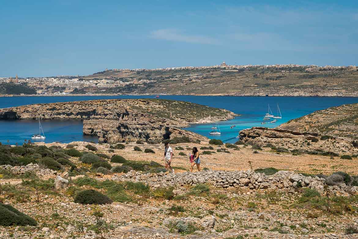 This image shows a couple hiking on a trail with the Blue Lagoon in the background.