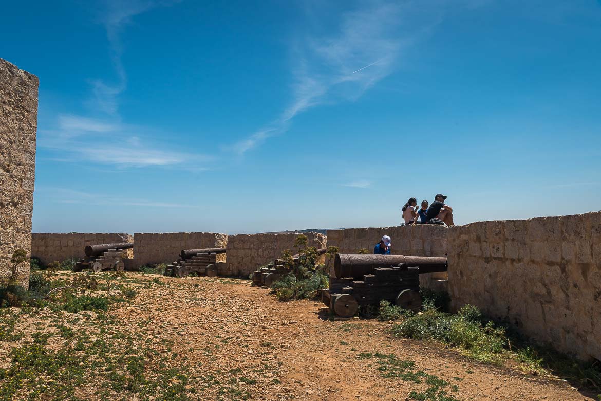 This image shows a family of four at Saint Mary's gun battery. Three of them are sitting on a high wall enjoying the sea view and the fourth one is standing next to one of the three cannons behind the wall.