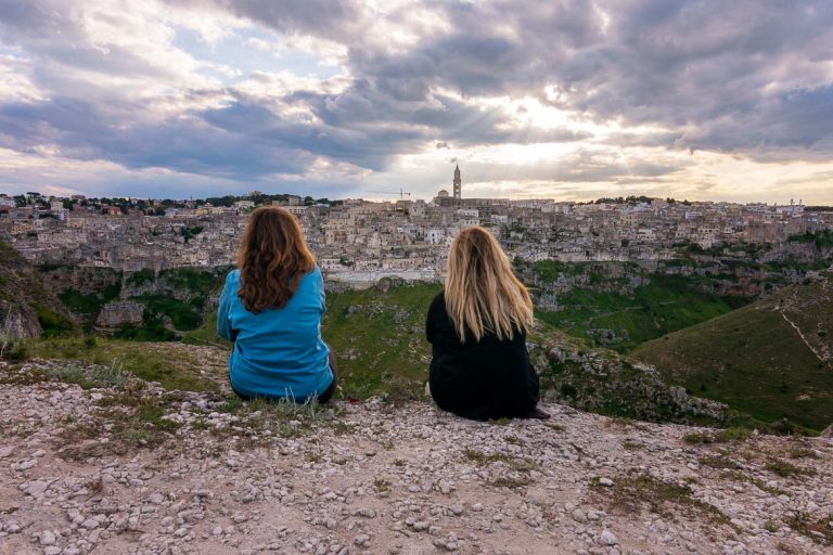 This photo shows Maria and Katerina with their backs turned to the camera sitting on a rock overlooking Matera. The Old Town looks amazing under a splendid sunset sky. This is the featured image of our article: How to travel more with a full time job: 8 tips to save time.
