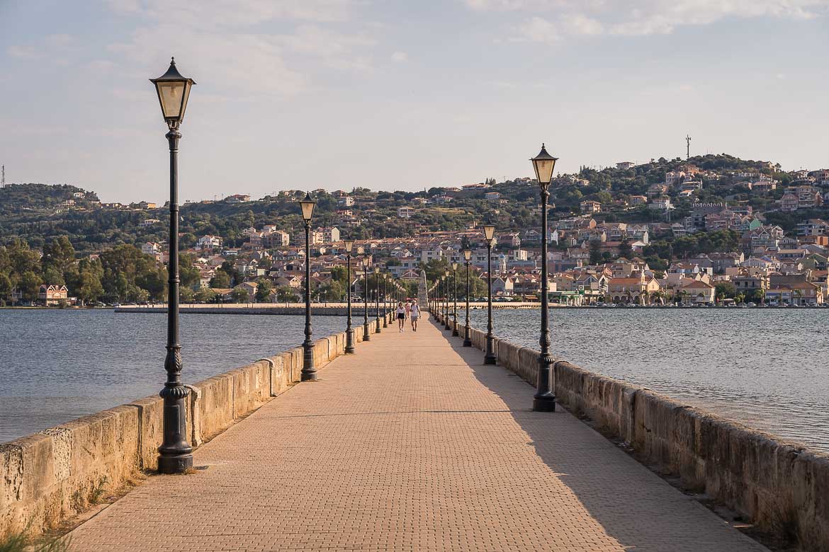 View of the De Bosset Bridge at sunset. The bridge is lined with street lamps and leads to Argostoli.