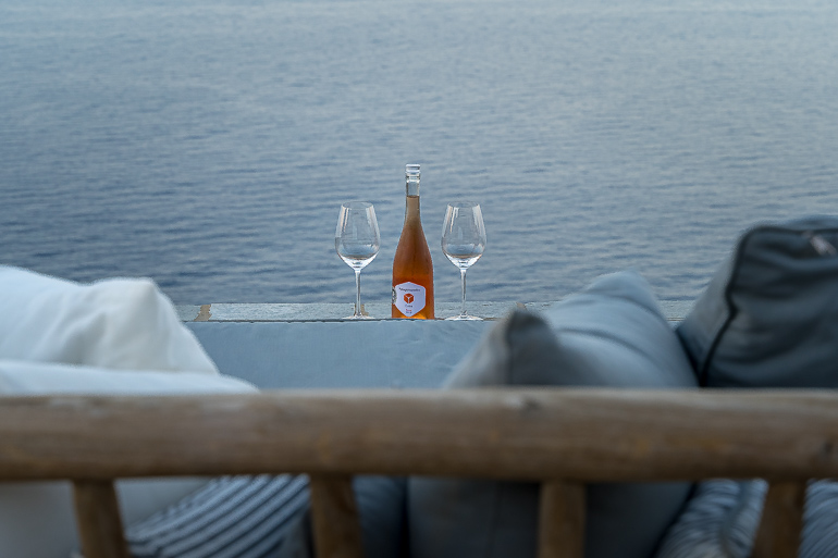 This image shows a bottle of wine with two glasses in the outdoor area of Verina Hotel in Sifnos.