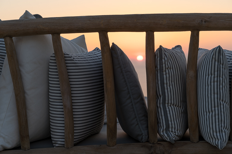 This image shows the sunset view through the pillows of a sofa from the outdoor area of Verina Hotel in Sifnos.