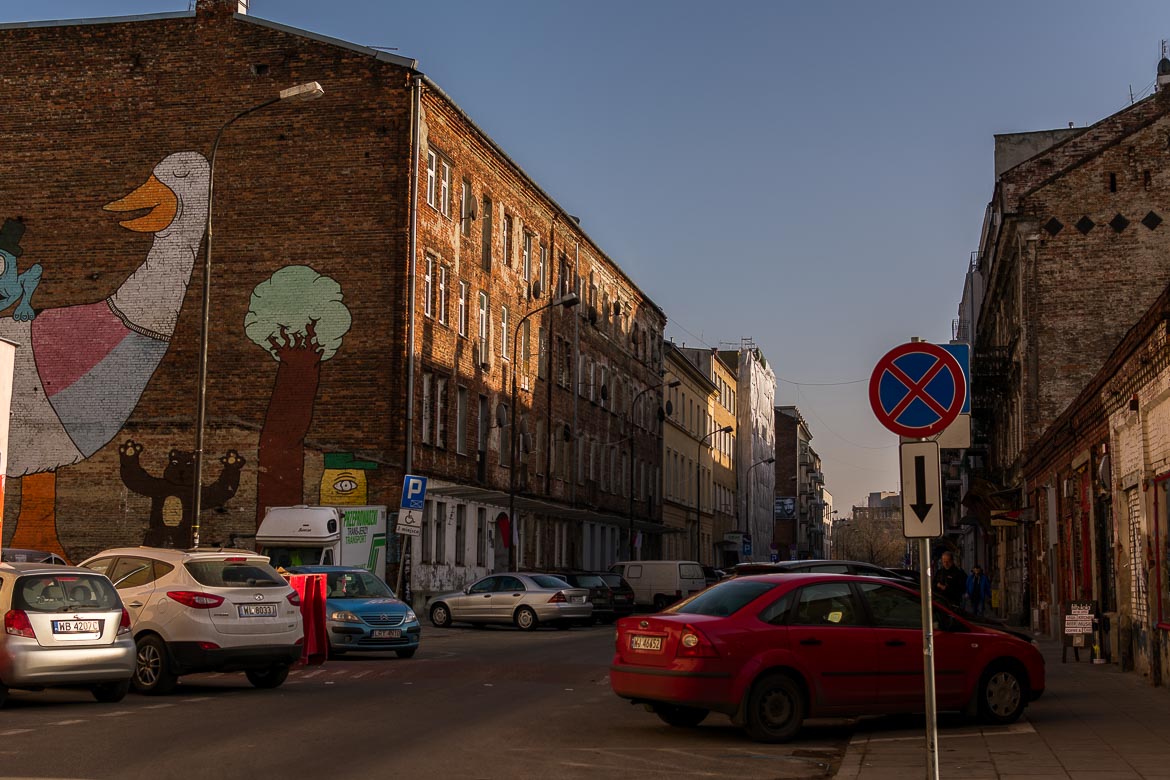 This image shows a street in the Praga district. There is a graffiti of a duck and a bear and several cars parked along the street. 