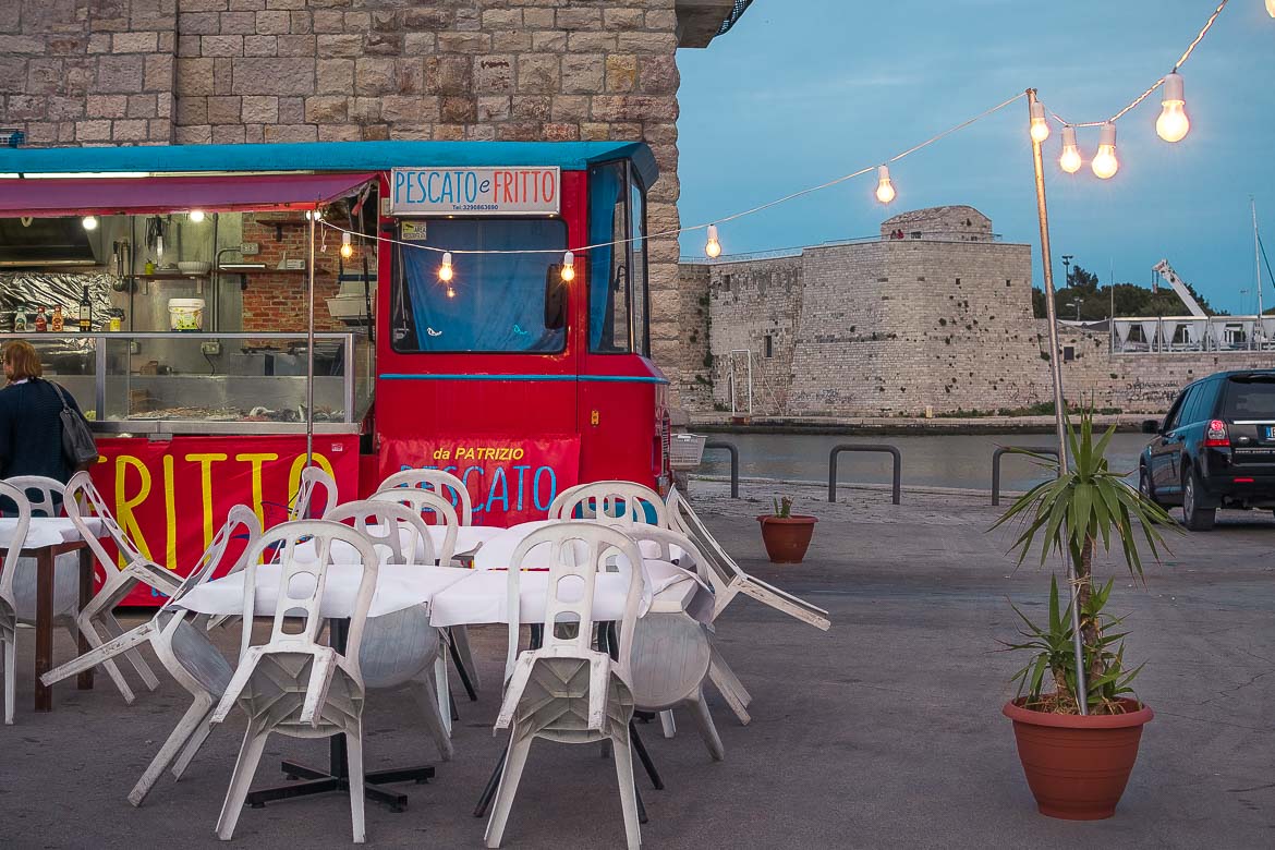 This photo shows a food truck on Trani promenade selling fried fish. The food truck is red and there is a line of lights hanging above. There are white tables and chairs. This photo is indicative of the simplicity of food in Puglia and this is why we chose it as the featured image for our Puglia food guide.
