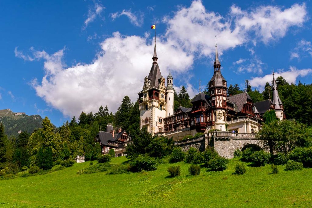 This is an image of Peles Castle. The castle looks magnificent with its elaborate towers and unique architecture. It is surrounded by splendid green landscape.