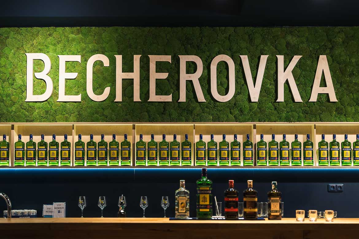 The bar of Jan Becher Museum. A big sign of Becherovka is over several shelves with bottles on the wall. Five bottles with different products of Becherovka and three shots are at the bar in the foreground.