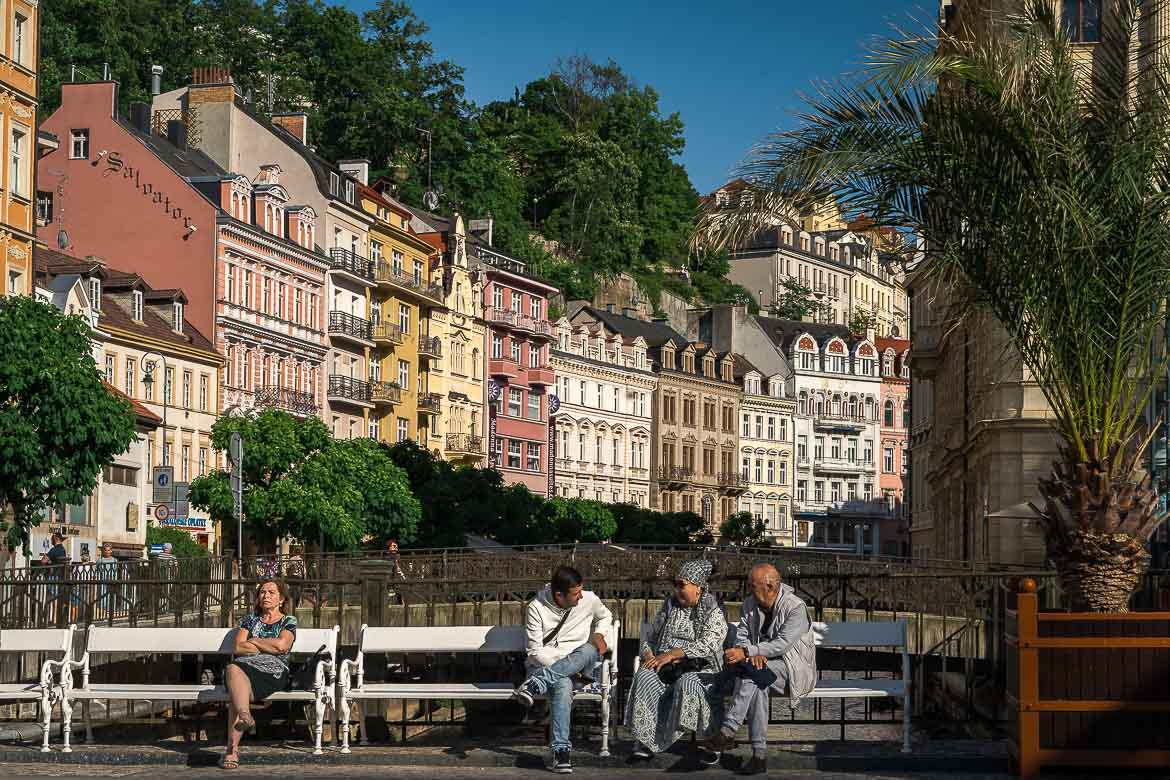 Four people are sitting on white benches in front of a bridge over the river. On the right, three of them are chatting while on the left a woman is sitting alone.
