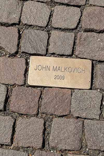 A golden cobble with John Malkovich 2009 written on it. The sign is placed on the pavement between other stones.
