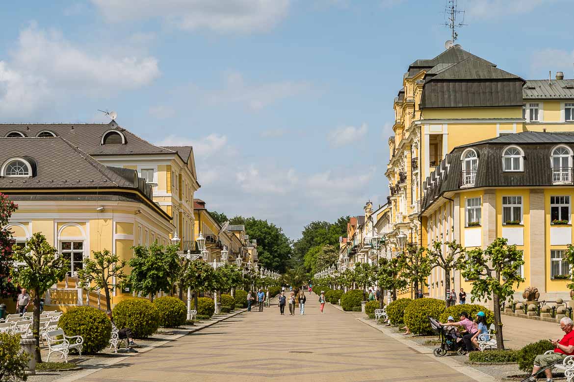 The main pedestrianised street in Frantiskovy Lazne. On both sides of the street, there are white benches, trees and bushes. The street is lined with elaborate yellow buildings with grey roofs.
