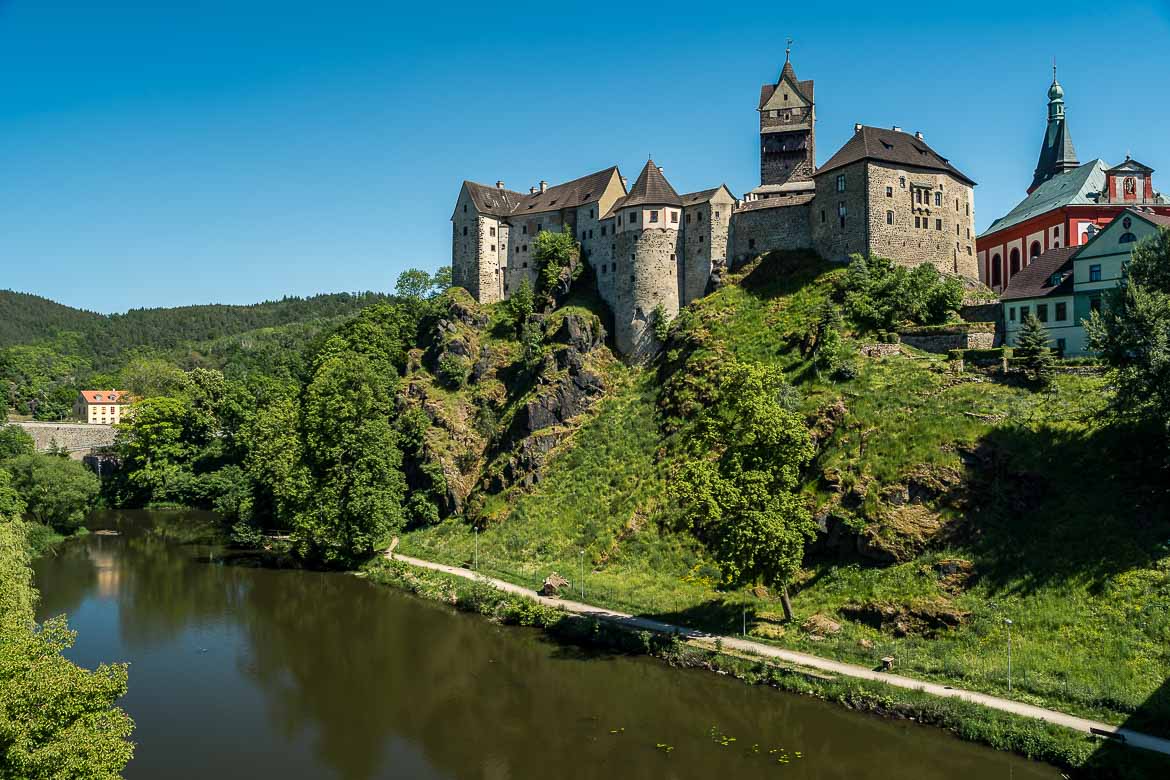Panoramic view of Loket Castle from the bridge. The castle is perched on a rocky hill. At the foot of the hill there is a river and a riverside promenade.