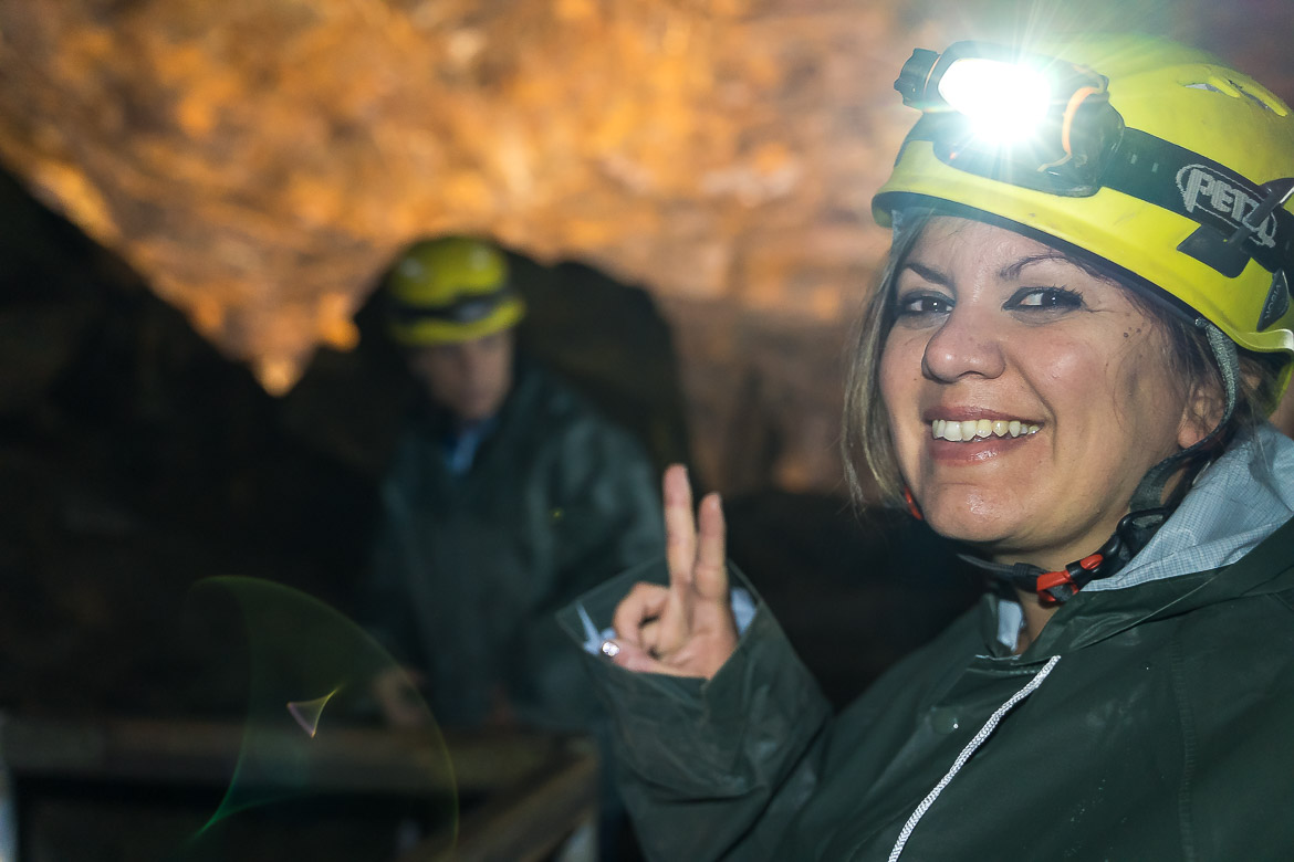 Maria wearing a yellow helmet with a light and a green anorak in the Johannes mine. She's doing the V sign at the camera, smiling. 