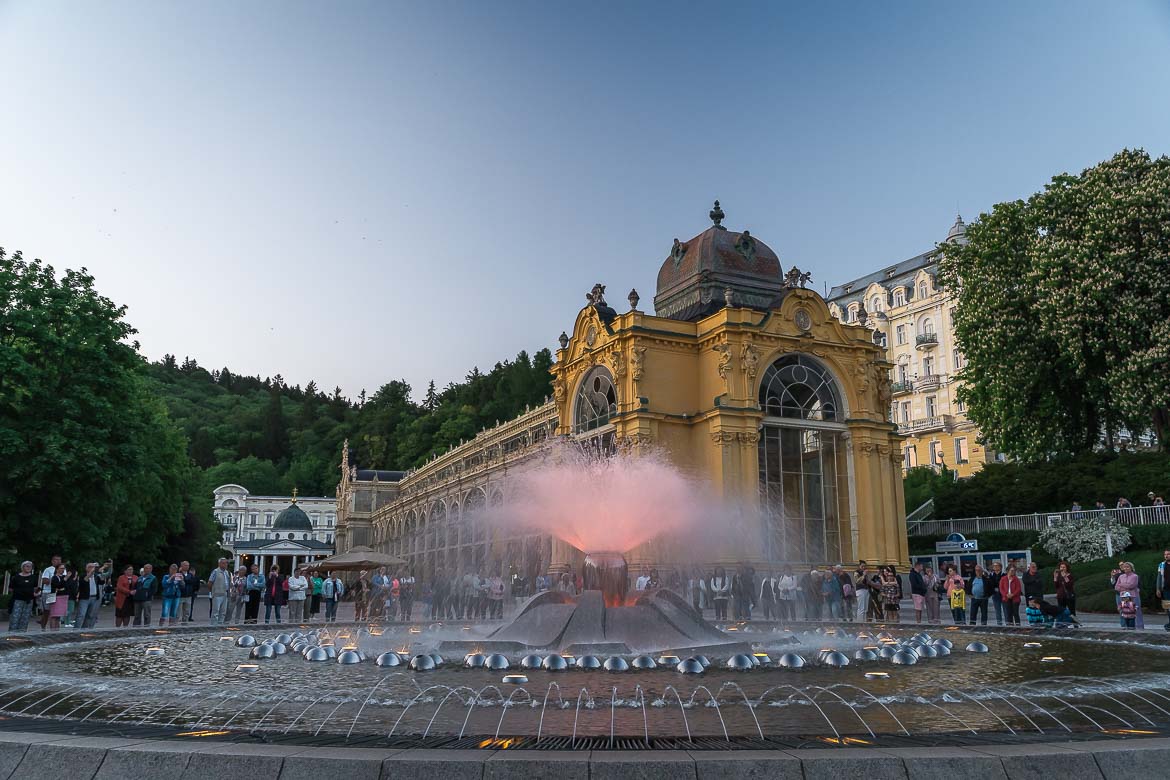 The Singing Fountain in Marianske Lazne during a music performance. The jet of the fountain has turned red and people are watching the show around the fountain. The Maxim Gorky Colonnade is in the background.
