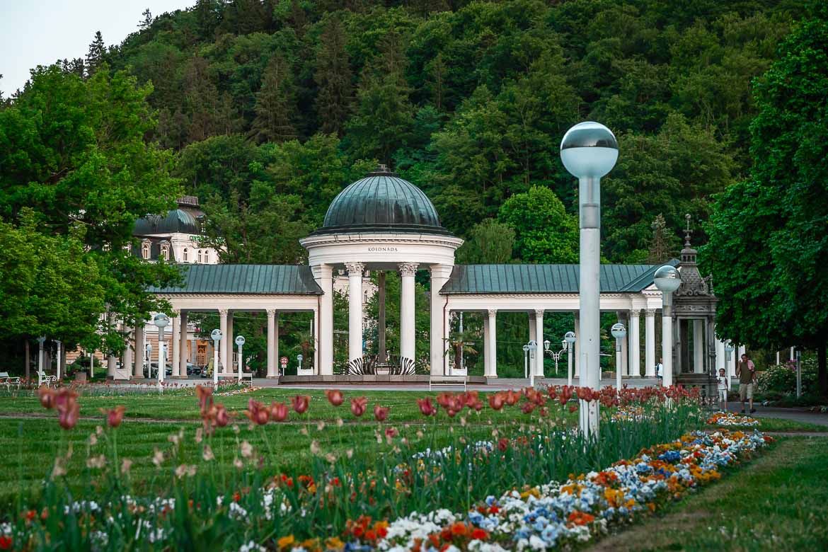 The colonnade with Karolina Spring. The colonnade is surrounded by a forest. Tulips and other flowers are in the background.