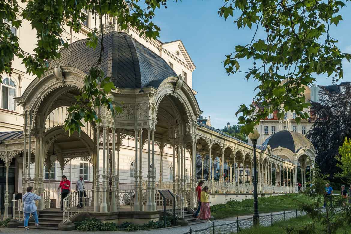 The iron-cast Park colonnade. It is formed by two gazebos linked with a corridor. People stand on the first gazebo in order to drink water from the spring and take photos. Walking along the colonnades is one of the best things to do in Karlovy Vary. 