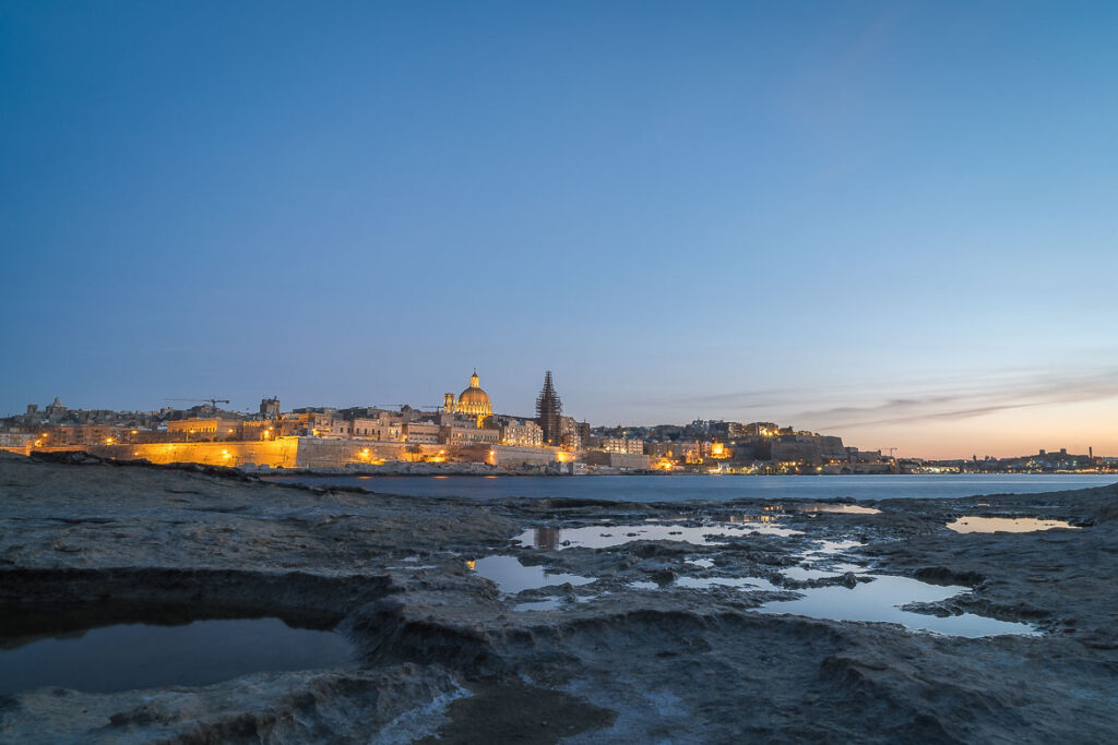 Panoramic view of Valletta from Tigne Point during the blue hour with pit holes filled with water in the foreground.