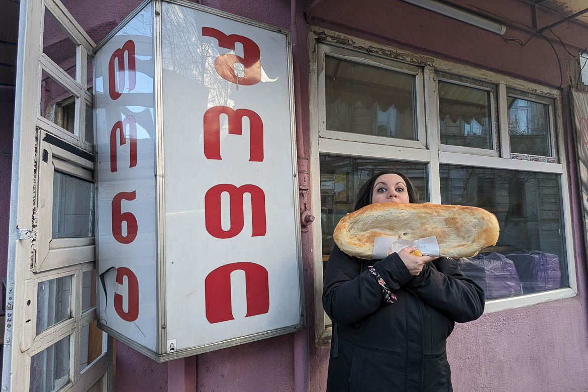 Maria holding a huge loaf of bread outside a traditional bakery. 