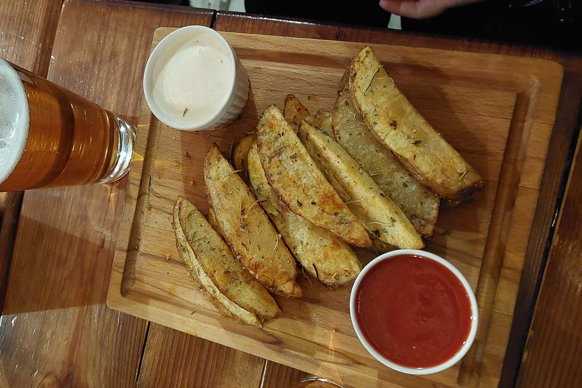 A close up of a wooden board with potato wedges topped with dried herbs. There are two small white bowls with dips, one red, one white. There's a glass of beer next to the board. 