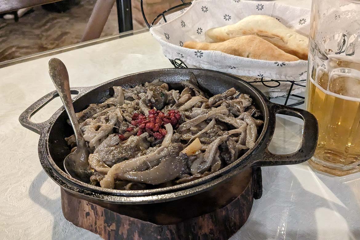 A close up of a cast iron pan with oyster mushrooms and a glass of beer next to it. In the background, there's a bread basket. 