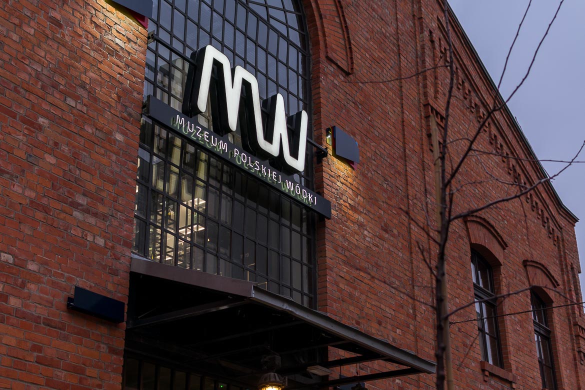 This image shows the facade of the Polish Vodka Museum. The facade is covered in red bricks and there's a neon sign with the museum's name. 