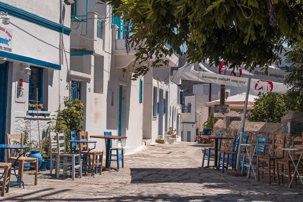 This image shows a cobbled street lined with whitewashed buildings and chairs and tables from two cafes.