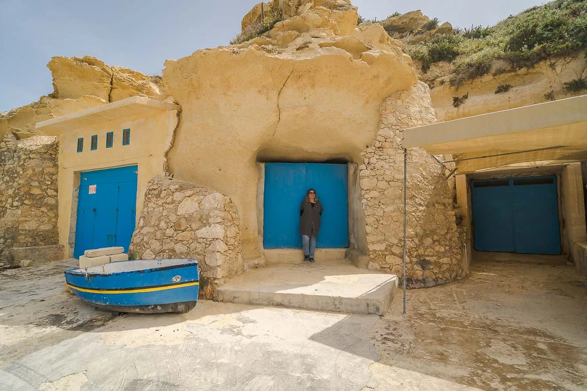 Maria is posing in front of a fisherman's shed. Thera are two more sheds. All three have blue doors. There is also a blue wooden boat next to one of the sheds.