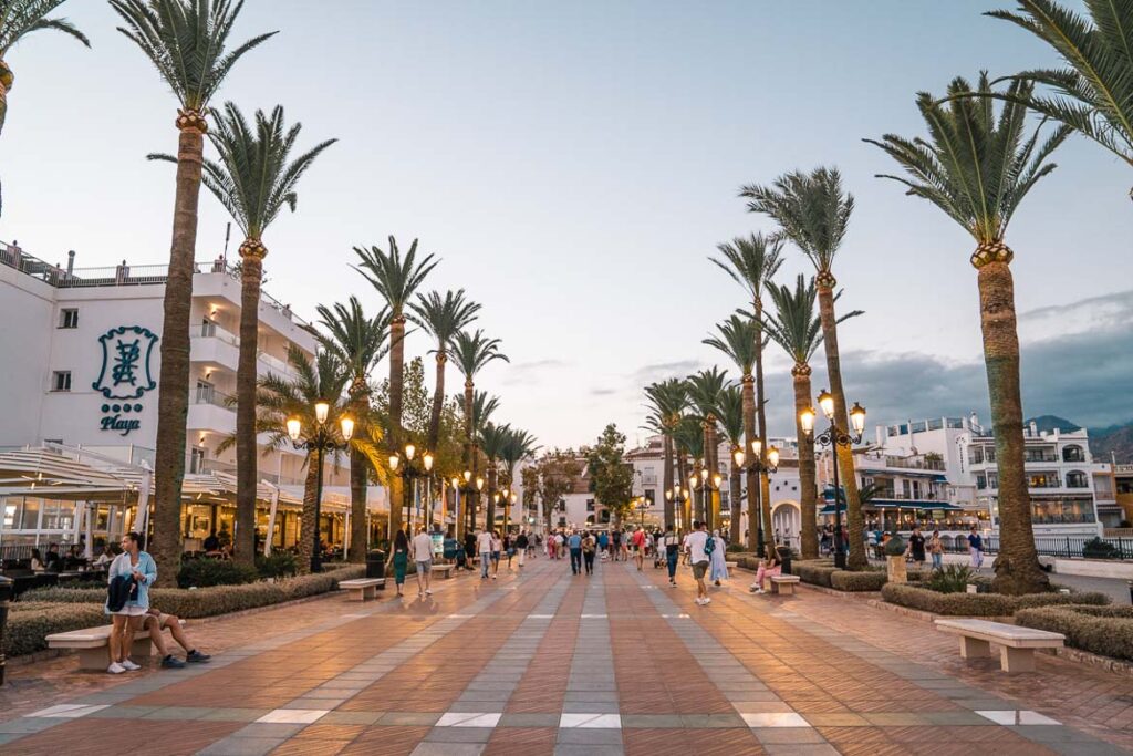 This image shows a wide pedestrianised boulevard lined by tall palm trees on both sides. It's dusk and the street lights are on. There are people walking along the boulevard. This is the featured image for our blog post What To Do in Nerja.