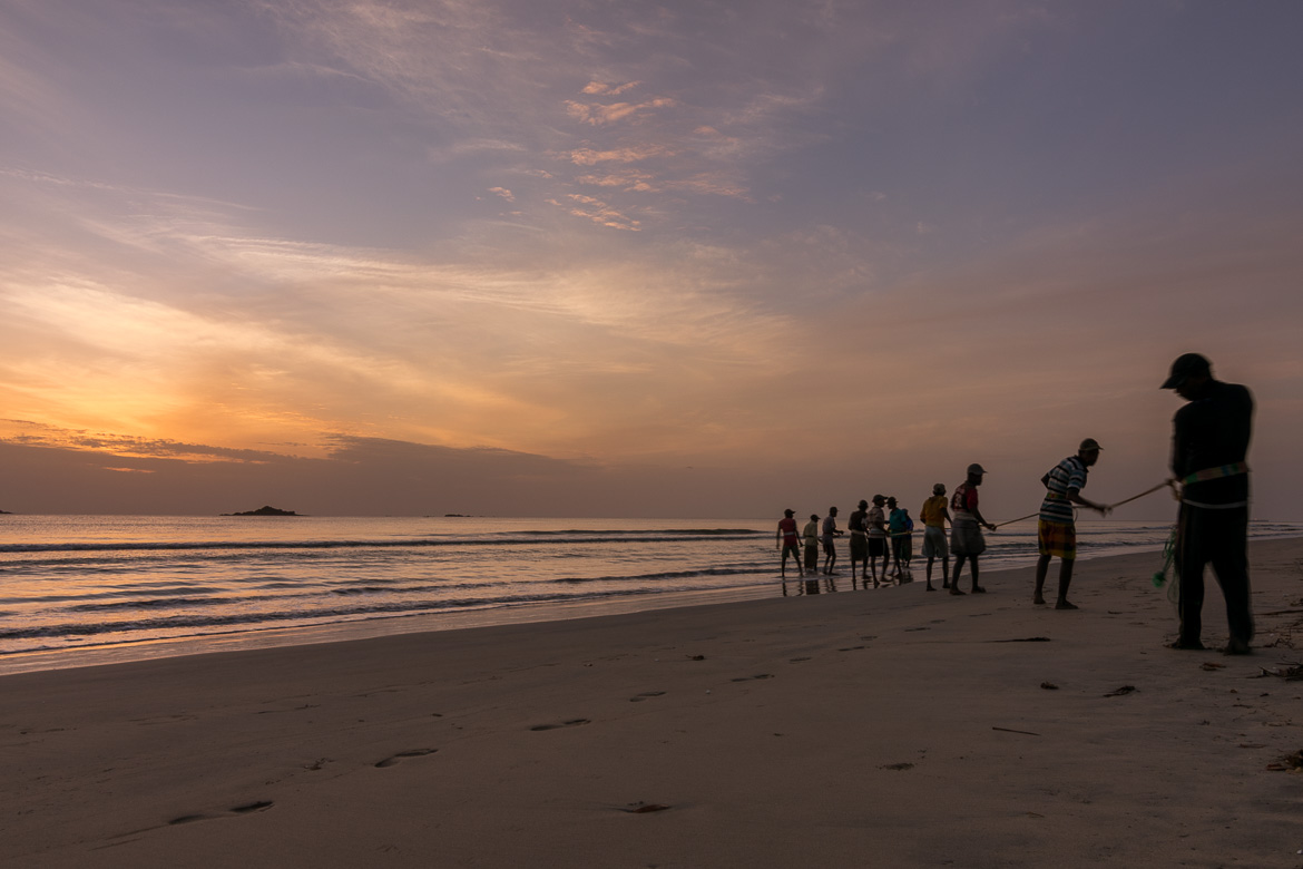 This photo shows fishermen on Nilaveli Beach pulling their nets from the sea at sunrise. The colour is a blend of blues, yellows and oranges. 